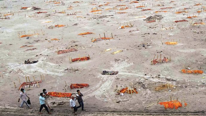 उथली कब्रें इलाहाबाद के उत्तर-पश्चिम शृंगवेरपुर में गंगा किनारे खुली कब्रों के बीच अपने रिश्तेदार का शव श्मशान ले जाते लोग, ये कब्रें कोविड मृतकों की बताई जाती हैं