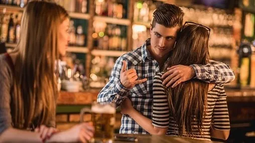 Unfaithful young man in a cafe. He is embracing his girlfriend and making call gesture to another woman behind her back.